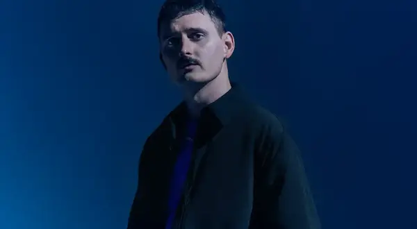 Dawid Podsiadło w ciemnej kurtce i fioletowej koszulce na niebieskim tle.