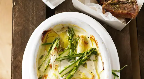 Überbackener Spargel mit Kartoffeln und Parmesan in einer Auflaufform, daneben ein Stück Lammkotelett.