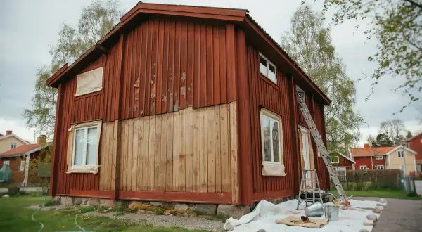 Ett rött trähus målas om. En stege står lutad mot huset, och färgburkar och penslar ligger utspridda på en presenning. Det ser ut som en perfekt dag att måla i solsken.