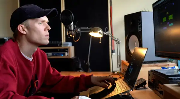 Ein junger Mann mit rotem Pullover und Kappe arbeitet an einem Laptop in einem Musikstudio. Er komponiert Musik, die die **lofi bedeutung** von Entspannung und Kreativität verkörpert.