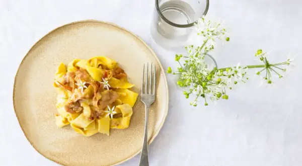 Ein Teller mit cremiger Pasta, die wie Fettuccine aussieht, garniert mit zarten Blüten und Speck. Daneben ein Glas Wasser und ein Strauß weißer Blumen.