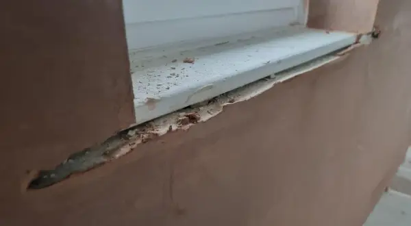 Fensterlaibung verputzen: Rauer Putz umrahmt ein weißes Fensterbrett, das mit kleinen Putzbrocken übersät ist.
