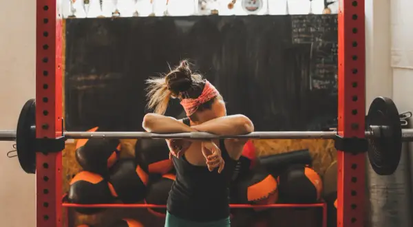 Erschöpfte Athletin lehnt an Hantelstange. Eine Woche Trainingspause ist sinnvoll für Regeneration und neue Motivation.