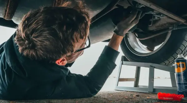 Mężczyzna w okularach, z rękawiczkami na dłoniach, pracuje pod samochodem. To jego kod zawodu mechanik samochodowy.