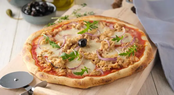 Une délicieuse pizza au thon, garnie de mozzarella fondante, d'oignons rouges et de roquette fraîche, prête à être découpée.