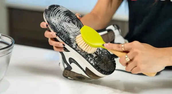 Main tenant une brosse verte, une personne nettoie la semelle extérieure d'une chaussure avec de la mousse.