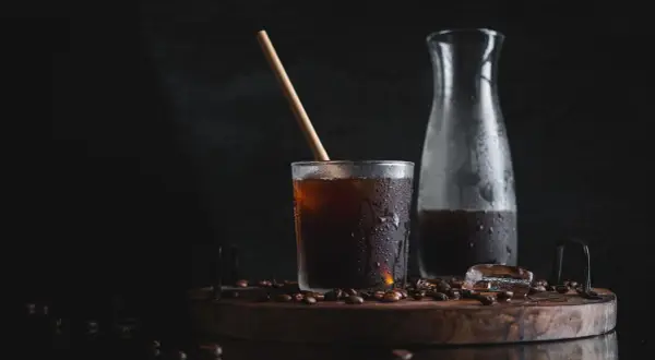 Ett glas med kallpressat kaffe, isbitar och en träpinne på en träbricka med kaffebönor. En karaff med kaffe står bredvid.
