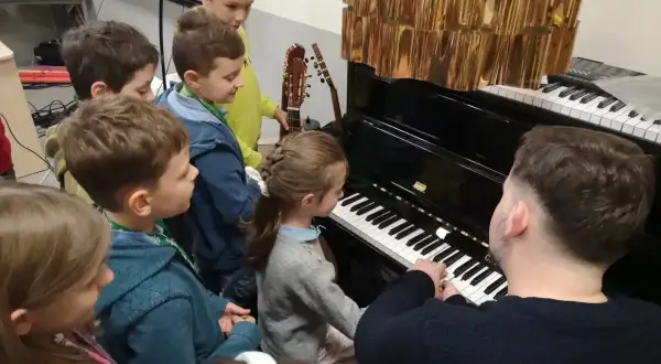 Dzieci słuchają lekcji muzyki przy pianinie. Nauczyciel pokazuje, do której klasy jest muzyka, a mali melomani z uwagą obserwują.