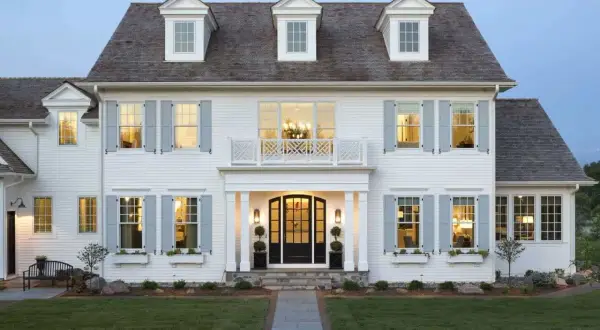 A stately white house with classic colonial architecture, featuring dormer windows, light blue shutters, and a welcoming porch.