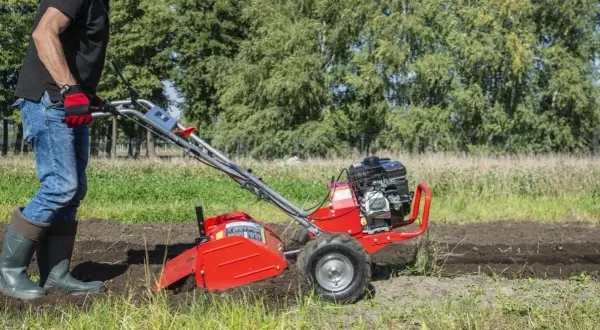 Mężczyzna w rękawiczkach i gumowcach obsługuje czerwoną glebogryzarkę na polu.
