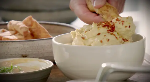 En hand doppar en bit i krämig hummus toppad med chiliflakes. Bredvid står en skål med gyllene halloumi fries.