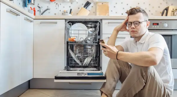 Zmartwiony mężczyzna szuka w telefonie informacji, jak zresetować zmywarkę Bosch, która stoi otwarta w kuchni.