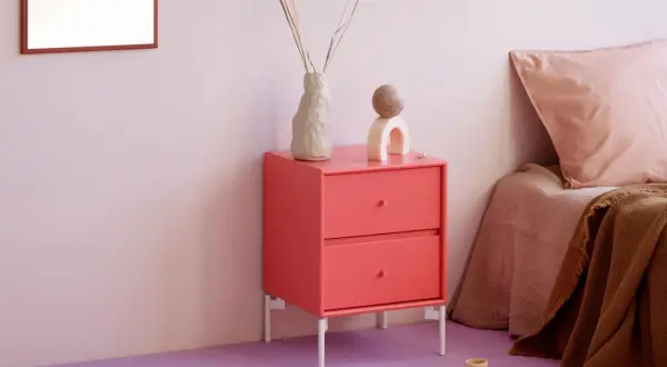 Ein Nachttisch in leuchtender **pink Farbe** steht neben einem Bett mit rosa Kissen. Dekorative Objekte und eine Vase mit Trockenblumen zieren die Oberfläche.