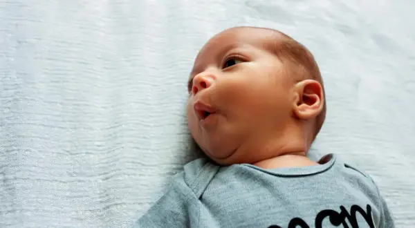 Uroczy niemowlak z kręczem szyi, ubrany w body z napisem "mom", leży na jasnoniebieskim tle.