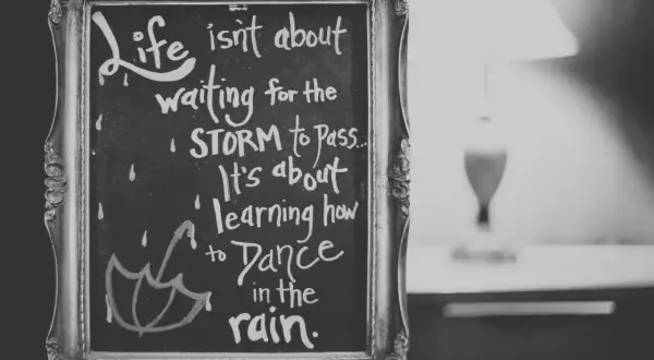 Czarno-biała tablica w ozdobnej ramie z inspirującymi cytatami po angielsku: "Life isn't about waiting for the storm to pass. It's about learning how to dance in the rain.