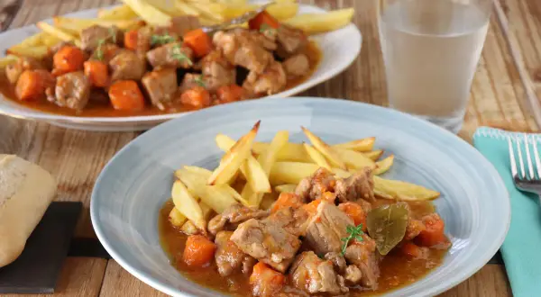 Dos platos de estofado de cerdo con patatas fritas y zanahorias, servidos en una mesa de madera.