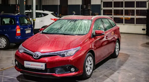 Czerwona Toyota Auris 1.6 benzyna z kroplami deszczu na karoserii, zaparkowana w garażu obok innych samochodów.