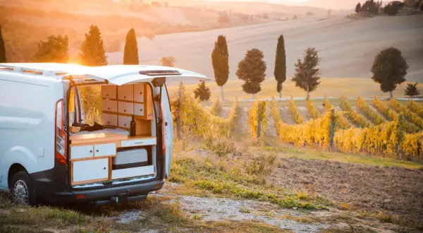 Un camper in un vigneto al tramonto, con dolci colline toscane sullo sfondo. L'interno del camper è visibile, con un letto e mobili.