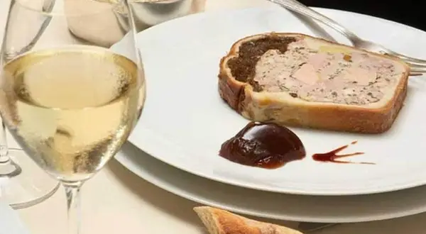 Une tranche de pâté en croûte, garnie de confiture de figues, accompagnée d'un verre de vin blanc et d'une coupelle de crudités.