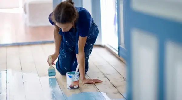 Mujer con mono azul se arrodilla para pintar el suelo de madera de azul claro. Se puede pintar el suelo para renovar la casa.