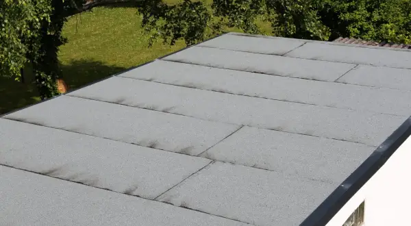 Graues Garagendach mit sichtbaren Rissen und Spalten, die auf ein undichtes Garagendach hindeuten. Im Hintergrund grüner Rasen und Bäume.