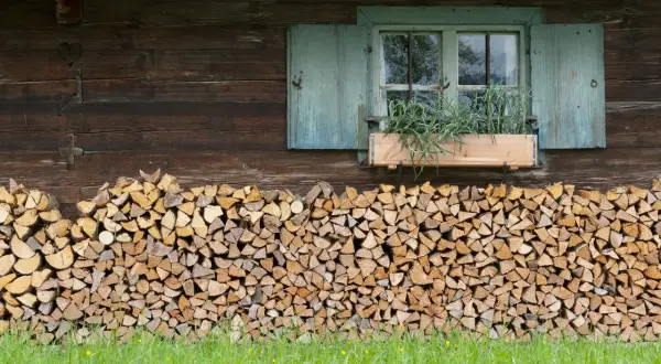 Ein ordentlicher Stapel Holz liegt vor der rustikalen Hauswand. Ein Fenster mit blauen Fensterläden und einem Blumenkasten mit Kräutern darüber.