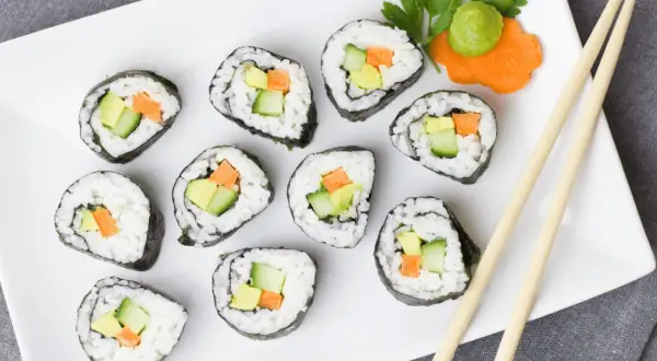 Jak ugotować ryż do sushi? Na talerzu ułożone rolki sushi z awokado, ogórkiem i marchewką. Obok pałeczki i dekoracja z wasabi.