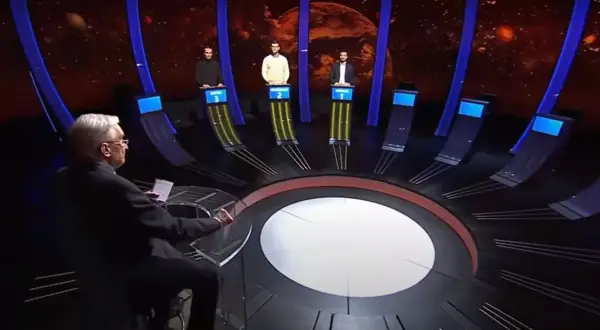 Trzech uczestników teleturnieju stoi przy swoich stanowiskach, a prowadzący siedzi przodem do nich.