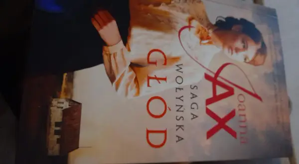 Okładka książki "Głód. Saga Wołyńska" Joanny Jax z portretem kobiety i budynkiem.