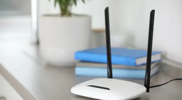 Biały router z dwiema antenami, zapewniający szybkie **standardy wifi**. W tle książki i roślina w doniczce.