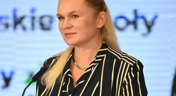 Kobieta z blond włosami w pasiastym ubraniu stoi przed mikrofonem na tle ekranu z tekstem.