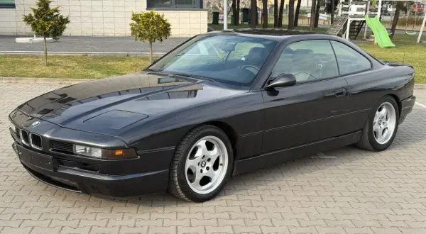 Czarne BMW V12, klasyk z lat 90., zaparkowane na kostce brukowej przed budynkiem z placem zabaw w tle.