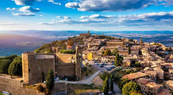 Scorci borghi antichi arroccati su una collina, con un castello imponente e case in pietra sotto un cielo azzurro con nuvole bianche.