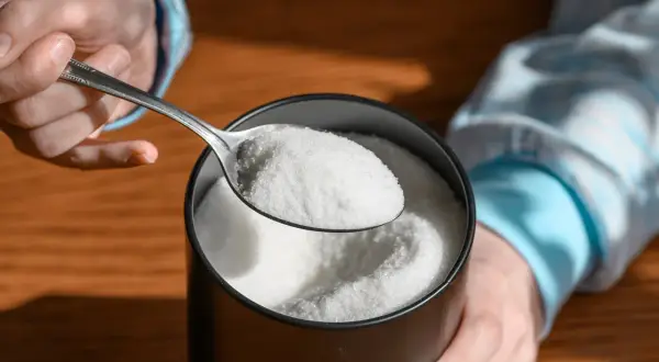 Mano che preleva eritritolo con un cucchiaio da un contenitore. Cos'è l'eritritolo? Un dolcificante a basso contenuto calorico.