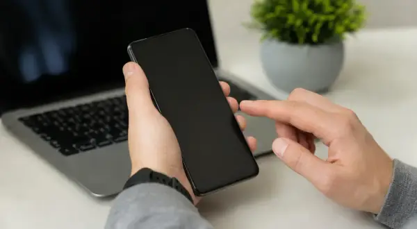 Dłoń trzyma smartfon, palec wskazujący dotyka ekranu, przygotowując się do całkowitego czyszczenia telefonu. Obok laptop i roślina.