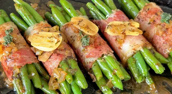 Grüne Bohnen im Speckmantel, mit Knoblauch und Kräutern verfeinert, liegen auf einer dunklen Platte in einer köstlichen Sauce.