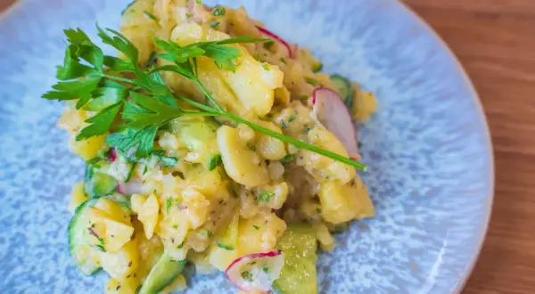 Ein köstlicher Kartoffelsalat mit Gurken, Radieschen und frischer Petersilie auf einem blauen Teller.
