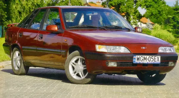 Czerwony Daewoo Espero z lat 90. na tle zieleni i brukowanej drogi.