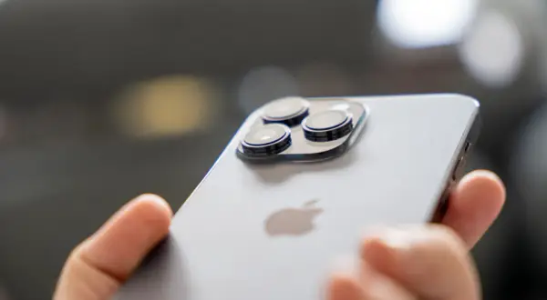 Dłoń trzyma iPhone 14 Pro, pokazując jego potrójny aparat. Długi czas pracy na baterii pozwala na swobodne korzystanie.