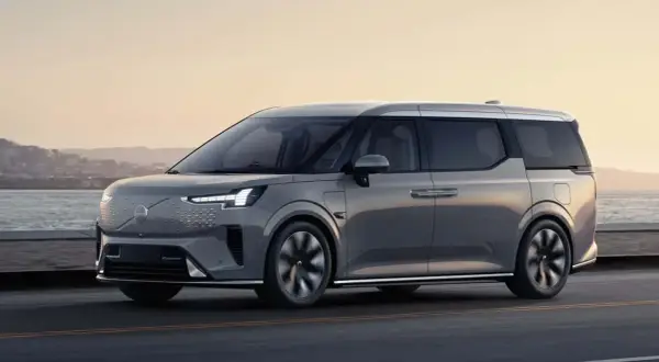 Elegancki, szary minivan Volvo EM90 jedzie wzdłuż wybrzeża o zachodzie słońca.