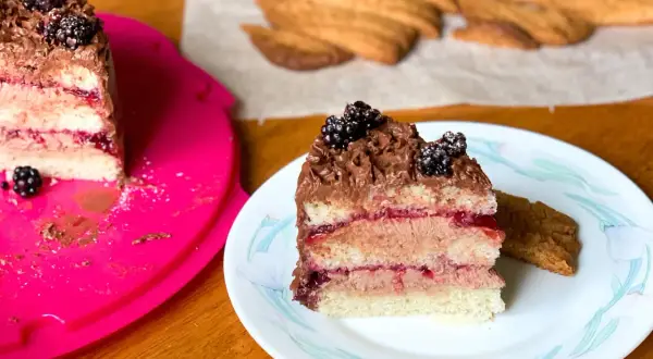 En bit av en läcker vegansk tårta med chokladfrosting och björnbär. Bredvid ligger en bit av en annan tårta och några kex.