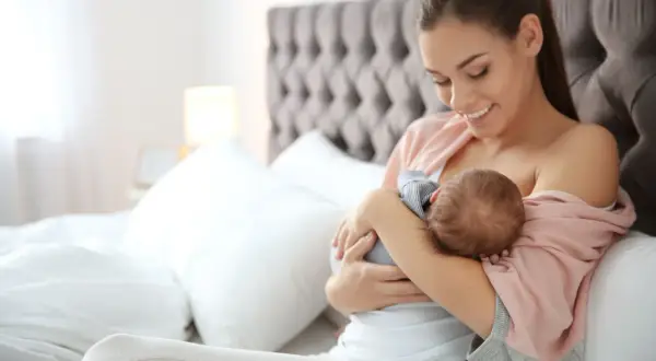 Uśmiechnięta mama karmi piersią noworodka. Czas karmienia jest elastyczny, ważne by dziecko było najedzone.