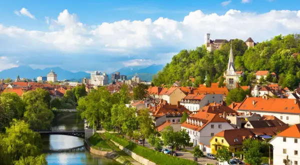Widok na rzekę Ljubljanicę, mosty i zabytkowe budynki Lublany ze wzgórzem zamkowym w tle.
