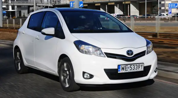 Białe małe auto, Toyota Yaris, jedzie po drodze. W tle budynki i torowisko tramwajowe.