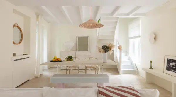 Un soggiorno luminoso con soffitto a travi e **bianco caldo pareti**. Tavolo da pranzo in legno, sedie, panca e una scala bianca conducono a un piano superiore.