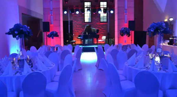 Elegancka filharmonia restauracja z fortepianem na scenie, stoły nakryte białymi obrusami i ozdobione kwiatami, oświetlone niebieskim i różowym światłem.