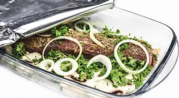 Mięso w naczyniu żaroodpornym, przykryte folią aluminiową, która pomaga utrzymać ciepło. Cebula i zioła dodają smaku.