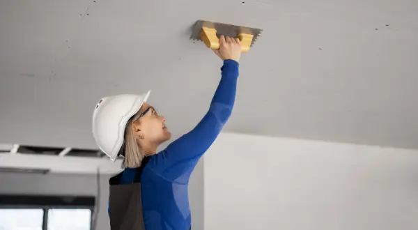 Kobieta w kasku i kombinezonie tynkuje sufit podwieszany kielnią.