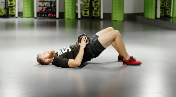 Mężczyzna z brodą wykonuje ćwiczenie glute bridge z obciążeniem na biodrach.