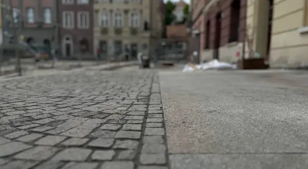 Nawierzchnia placu miejskiego, gdzie kostka brukowa sąsiaduje z betonem. W tle widać budynki i ślady prac remontowych, symbolizujące postępującą betonoza.